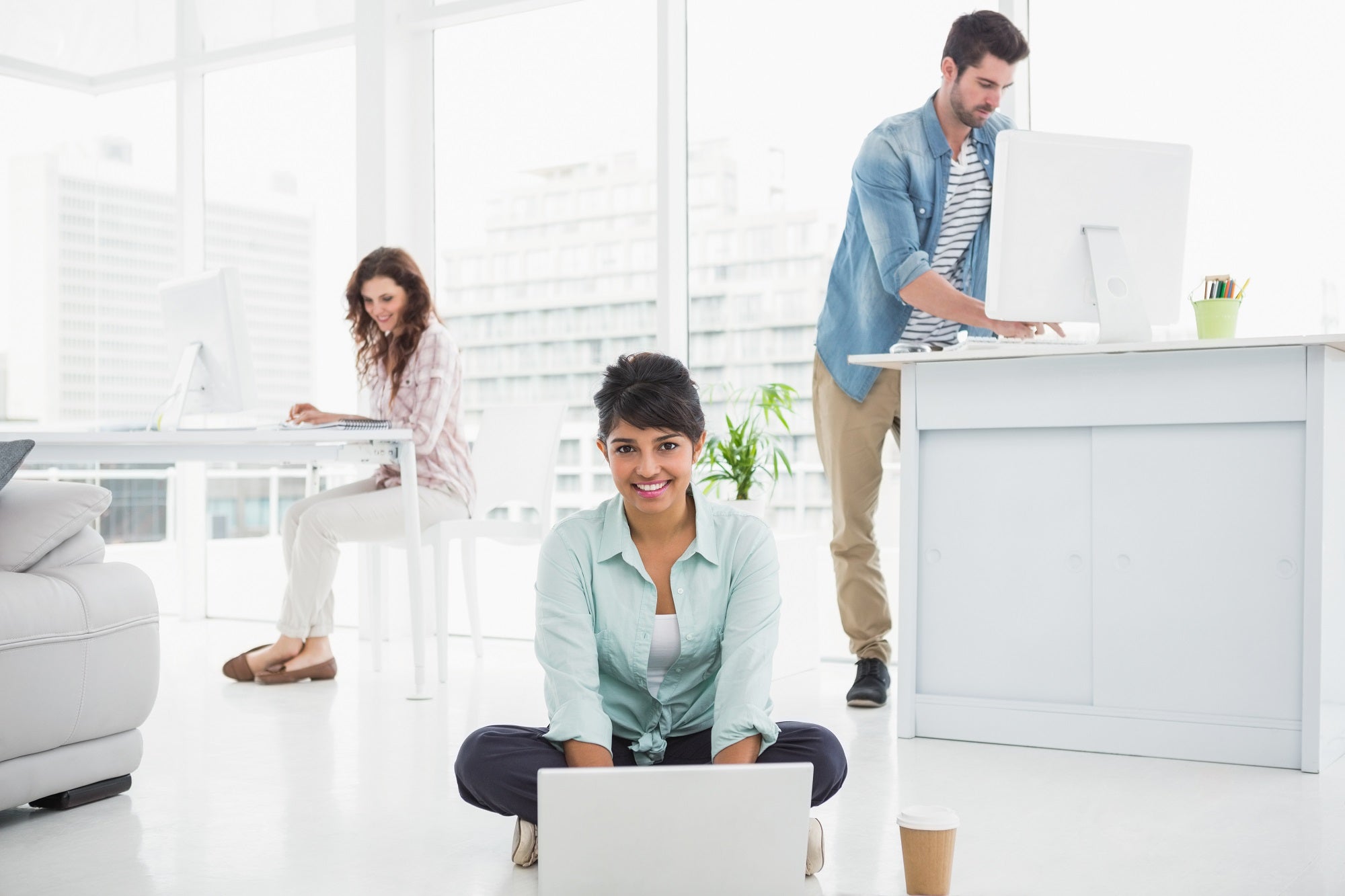 Smiling businesswoman sitting on the floor using laptop with colleagues behind her